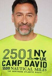 Camiseta de manga corta de color verde lima brillante con texto gráfico negro que incluye "CAMP DAVID" y detalles de temática náutica.