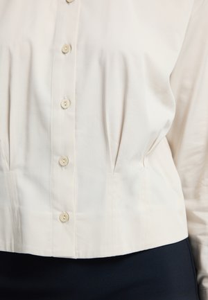 Cream blouse with five buttons, featuring pleats and a structured fit. Made from smooth fabric, it has a rounded hem and long sleeves.