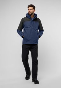 Veste imperméable bleue avec un panneau d'épaule noir, capuche ajustable, fermeture éclair à l'avant et poches latérales, assortie avec un pantalon noir et des bottes texturées.