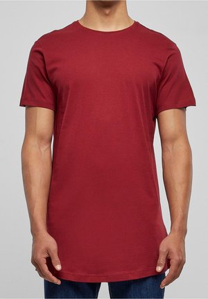 Uomo che indossa una semplice maglietta rossa a maniche corte con colletto tondo e jeans blu, in piedi contro uno sfondo grigio, torso visibile.