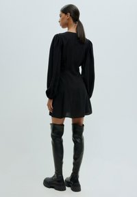 Schwarzes kurzes Kleid mit Puffärmeln und taillierter Passform. Getragen mit hohen schwarzen Lederstiefeln mit dicker Sohle. Glatte Textur, minimalistisches Design.