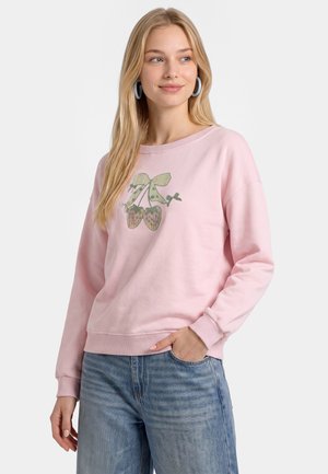 Ung kvinde med langt blondt hår iført en lyserød sweatshirt med kirsebærdesign og blå jeans, smilende og kigger til siden.