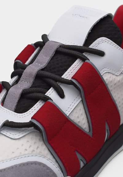 Κοντινό πλάνο ενός αθλητικού παπουτσιού με μαύρα κορδόνια, άσπρο και μαύρο δίχτυ, γκρι σουέτ λεπτομέρειες και μεγάλο κόκκινο λογότυπο «N» από σουέτ στο πλευρό.