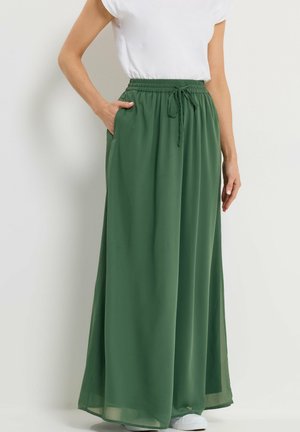 Nainen, jolla on päällään pitkä, vihreä, liehuva hame, jossa on joustava vyötärönauha ja kiristysnauha, yhdistettynä valkoiseen t-paitaan ja valkoisiin tennareihin.