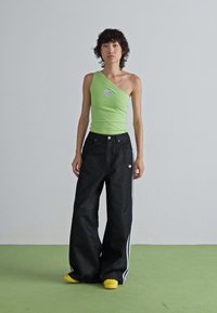 Jonge vrouw staat op een groene vloer en draagt een limoen groene one-shoulder top met een strass hart, zwarte wijde broek en gele schoenen.
