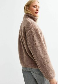 Bruine fleece jas met hoge kraag, boxy fit en een gestructureerd oppervlak. De mouwen zijn oversized en het is gecombineerd met lichtgekleurde spijkerbroeken.
