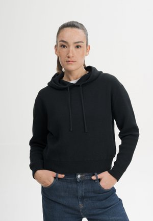 Vrouw met naar achteren getrokken haar, gekleed in een zwarte hoodie en blauwe spijkerbroek, staat met handen in de zakken tegen een effen witte achtergrond.