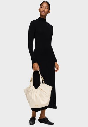 Donna che indossa un lungo abito nero con collo alto, tiene una grande borsa beige e porta scarpe nere piatte con punta, in piedi su sfondo bianco.