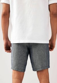 Shorts en denim foncé avec une finition texturée, dotés de revers roulés et de poches latérales, portés sous une chemise blanche légère et ample.