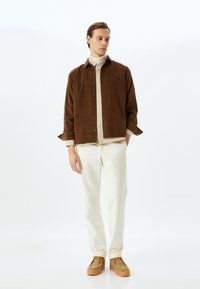 Chaqueta de botonadura en pana marrón sobre una camisa beige, combinada con pantalones de pana crema y calzado marrón. Texturas suaves y diseño en capas.