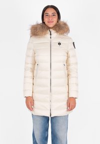 Crème pufferjas met een nep bontkap, horizontale naden, ritszakken en logo-detail op de borst. Gemaakt boven lichte jeans.