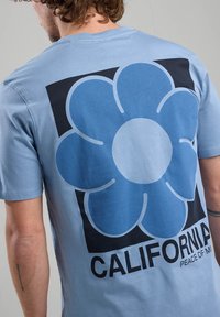 Vaaleansininen puuvillapaita, jossa on suuri sininen kukkakuva mustalla neliötaustalla, ja alla teksti "CALIFORNIA PEACE OF MIND".