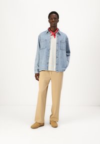 Τζιν πουκάμισο σε ανοιχτό μπλε, κρεμ πουλόβερ από κάτω, μπεζ παντελόνι με φαρδιές γάμπες και καφέ αθλητικά παπούτσια. Η κόκκινη μπαφ να δίνει μία ζωντάνια χρώματος.