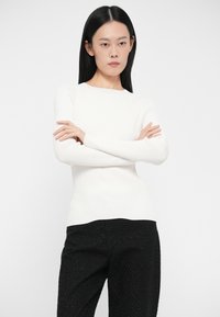 Camisola branca de mangas compridas com decote arredondado, design ajustado, combinada com calças pretas texturizadas com pequenos pontos.