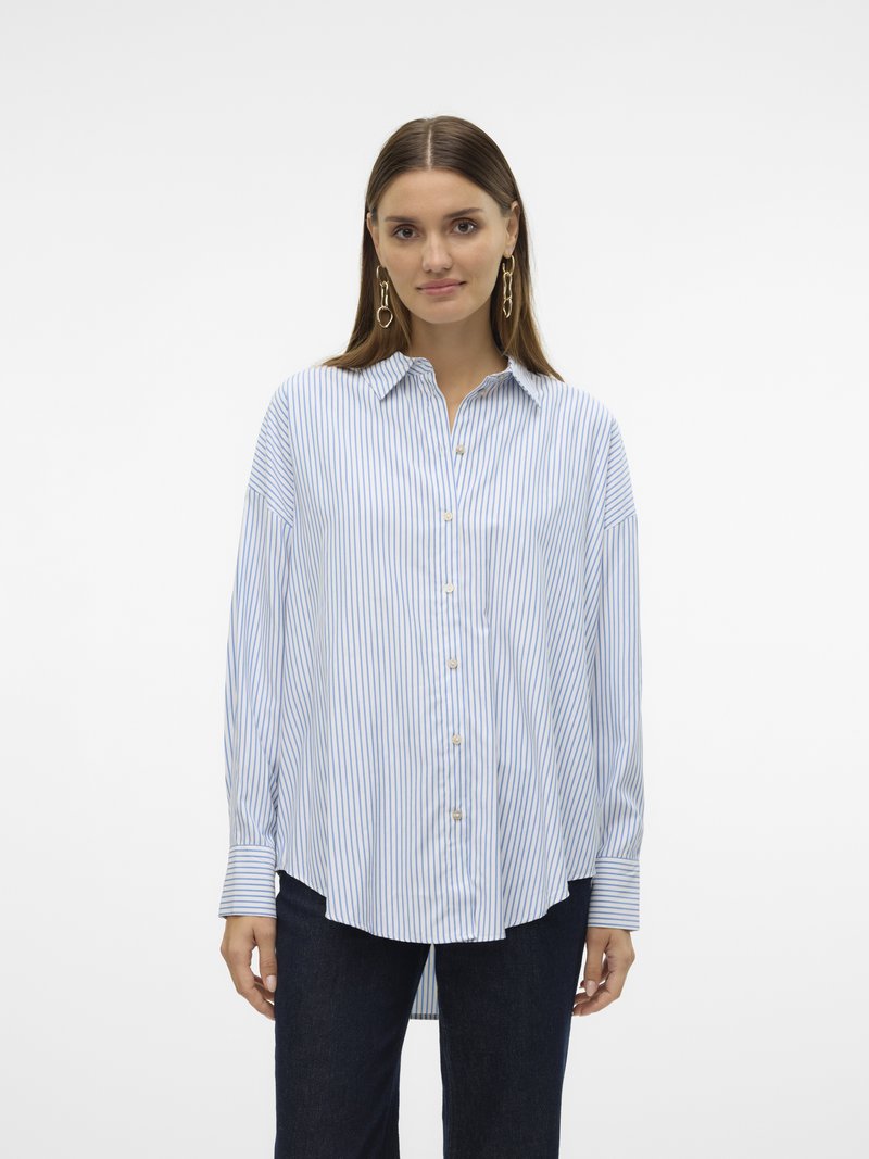 Kvinne iført en løs, langermet, lyseblå og hvit vertikaltstripet skjorte med mørke bukser og gullkjede-øredobber.