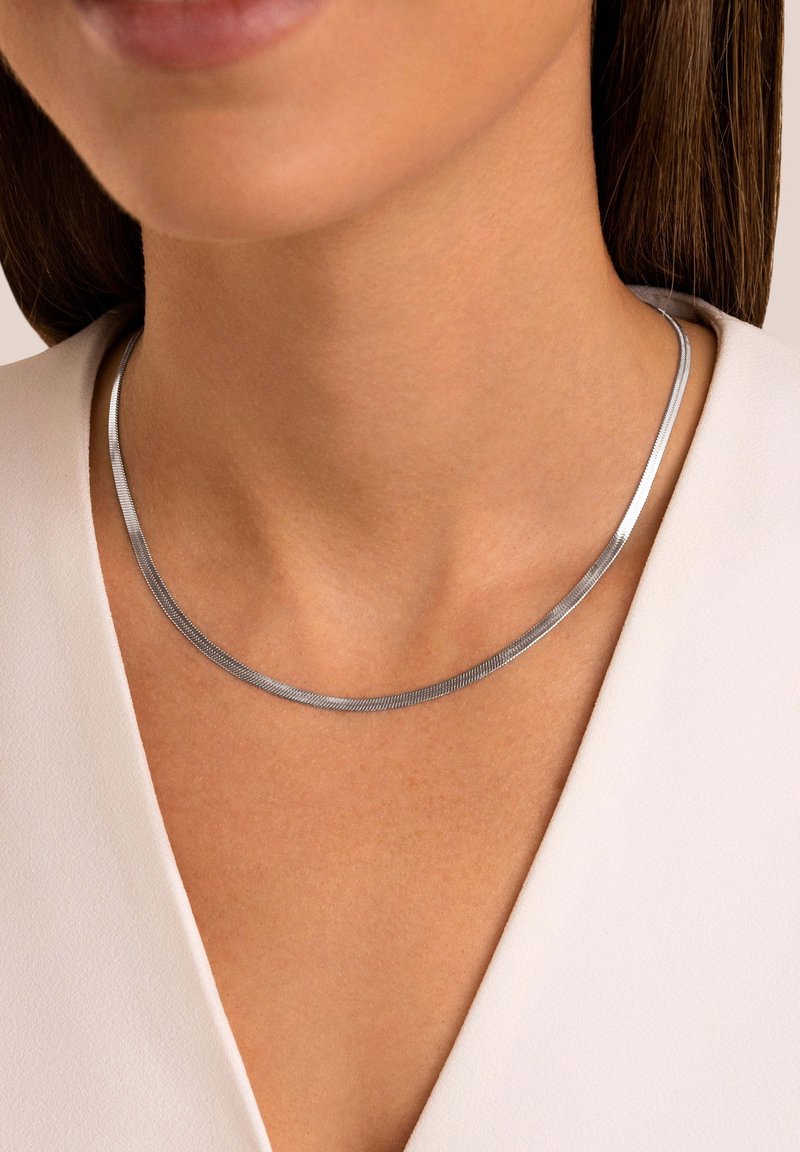 Gros plan d'une personne portant un collier chaîne serpent lisse argenté avec un haut blanc à col en V et des cheveux bruns raides.