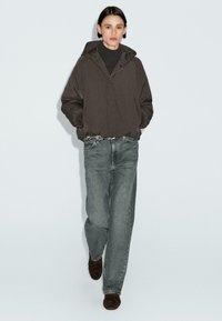 Donna che cammina indossando un giubbotto con cappuccio scuro, un maglione nero, jeans grigi larghi con una cintura borchiata e scarpe marroni scure su uno sfondo semplice.