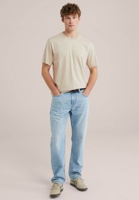 Béžové tričko s krátkým rukávem, volný střih, v kombinaci se světle modrými džíny. Outfit doplňují béžové a zelené tenisky. Jednoduché pozadí.