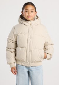 Un giovane che indossa una giacca a vento beige con cappuccio e jeans azzurri, in piedi con una mano in tasca su uno sfondo neutro.
