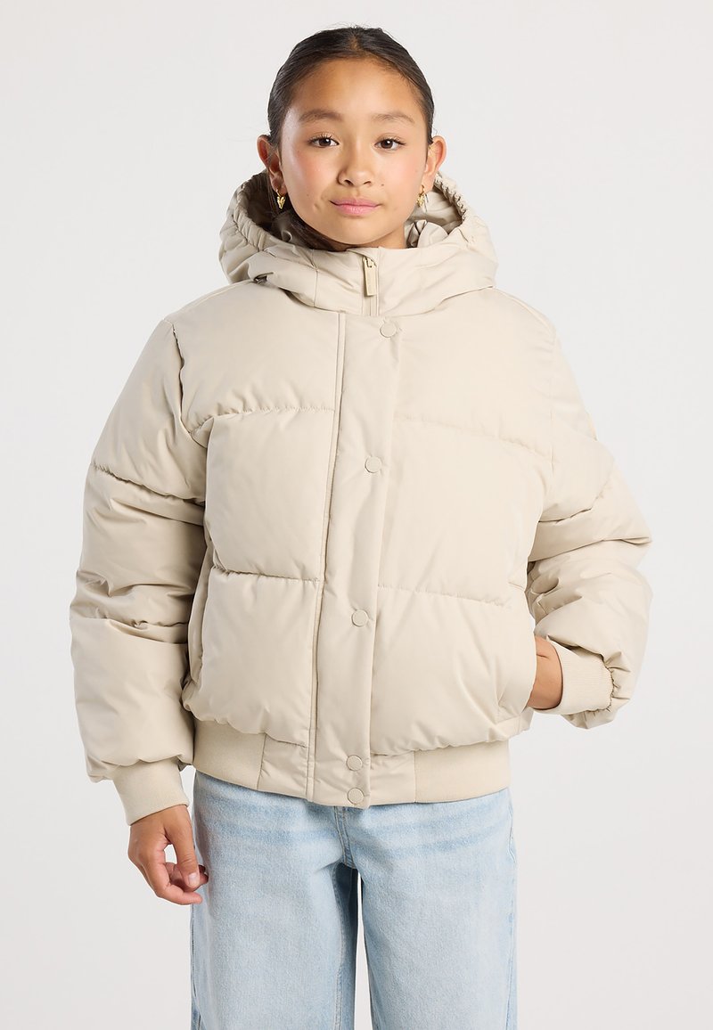 Junge Person trägt beige Steppjacke mit Kapuze und hellblaue Jeans, steht mit einer Hand in der Tasche vor einfarbigem Hintergrund.