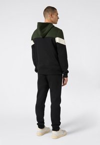 Uomo che indossa un felpa nera e verde scuro con strisce bianche sulle maniche, pantaloni della tuta neri e scarpe da tennis bianche, in piedi di spalle.