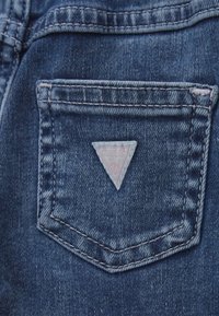 Denim tkanina, tamnoplave boje, mali stražnji džep s trokutastim Guess logom u svijetloružičastoj boji, obrubljen bijelim šavom.