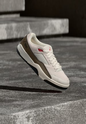 Une basket blanche et marron avec des accents rouges flotte au-dessus de marches en béton, montrant son profil latéral et ses lacets à la lumière du jour.