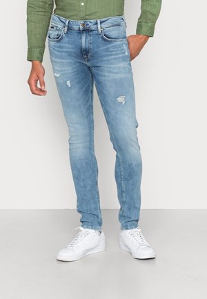 Mand iført lyseblå slim-fit revnede jeans, hvide sneakers og en langærmet grøn skjorte med knapper, stående mod en ensfarvet baggrund.