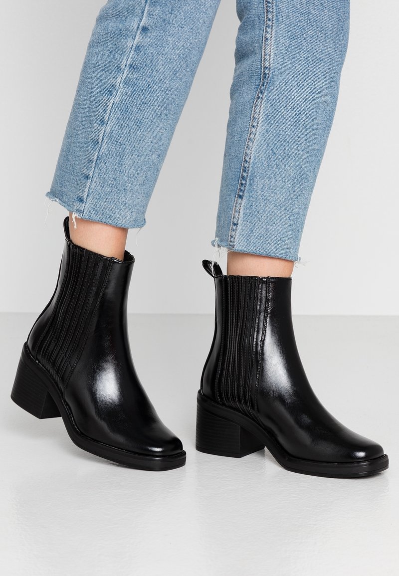 Persona vistiendo jeans cortos de color azul claro y botas de cuero negro brillantes con tacones cuadrados sobre un suelo blanco.