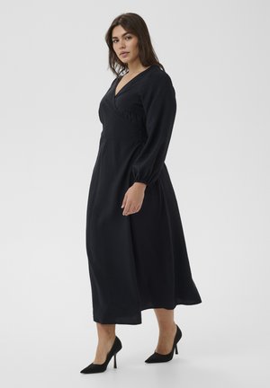 Frau steht in einem langen schwarzen Kleid mit langen Ärmeln und schwarzen High Heels vor einem schlichten hellen Hintergrund.