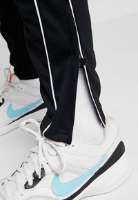 Zwarte sportbroek met witte strepen aan de zijkant en ritsen bij de enkels. Draagt witte sneakers met accenten in lichtblauw en zwart.