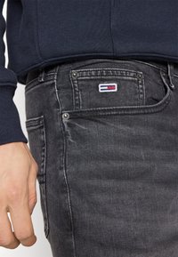 Prim-plan al unei perechi de blugi din denim gri închis, având un mic logo dreptunghiular pe buzunarul pentru monede, lângă o mână și tivul unui hanorac de culoare bleumarin.