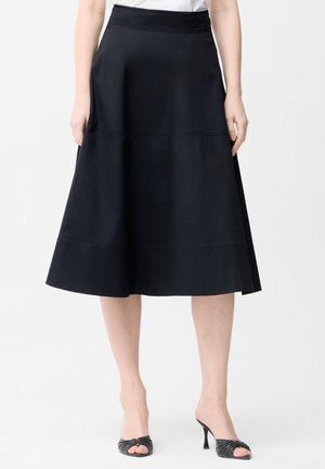 Frau trägt einen knielangen schwarzen Rock im A-Linien-Stil und schwarze offene High-Heels, steht vor einem einfarbigen weißen Hintergrund.