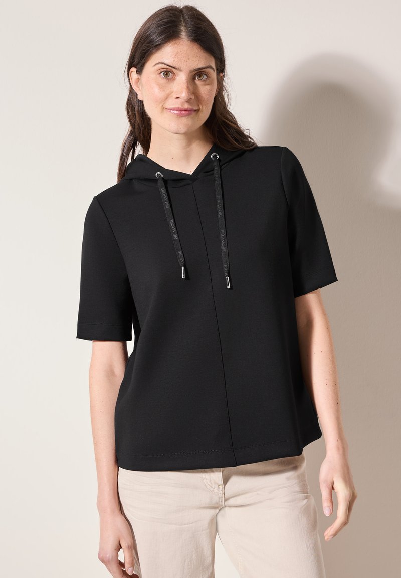 Sudadera corta negra con manga corta y cremallera frontal, con cordones con puntas metálicas. La tela es suave y ligeramente elástica.