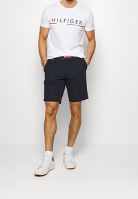 Bílý bavlněný tričko s logem "HILFIGER", doplněné námořnickými kraťasy. Model nosí bílé tenisky s námořnickými akcenty a ponožky.
