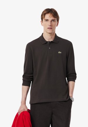 Čierna polo košeľa s dlhým rukávom a golierom, so zeleným logom. Model drží červenú bundu a stojí na bielom pozadí.