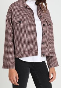 Giacca a scacchi cropped in bordeaux e crema, con due tasche sul petto, bottoni neri e un fit rilassato, abbinata a un dolcevita bianco.