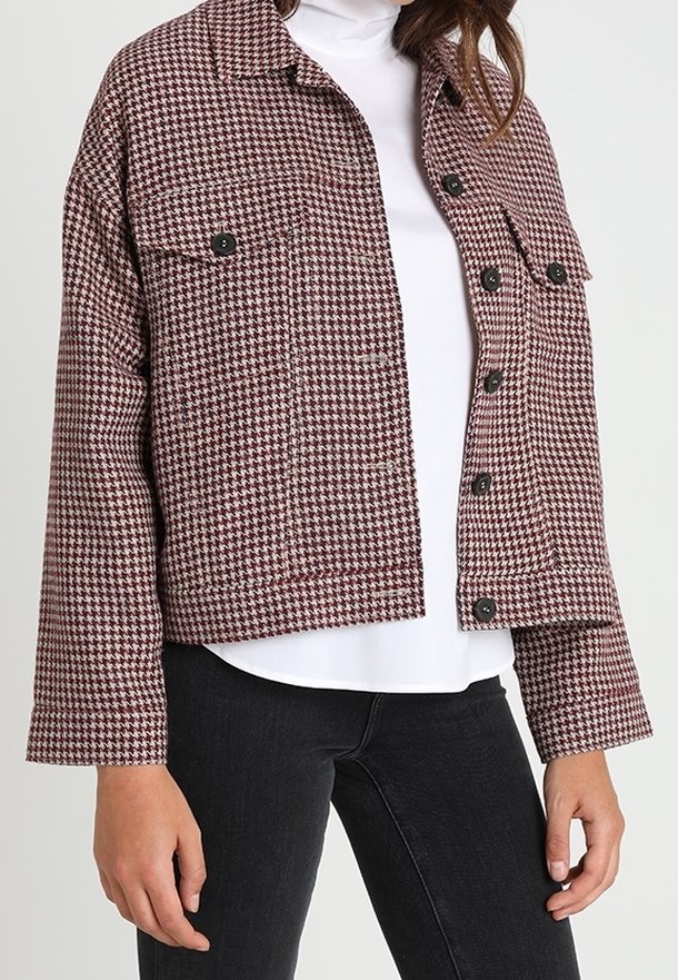 Giacca a scacchi cropped in bordeaux e crema, con due tasche sul petto, bottoni neri e un fit rilassato, abbinata a un dolcevita bianco.