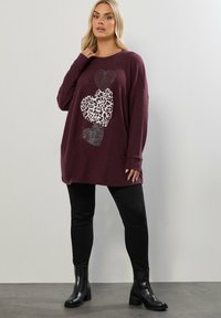Bordeaux oversized tröja med hjärtan i olika texturer, inklusive leopardmönster och zebraränder, kombinerad med svarta byxor och stövlar.