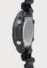 Montre numérique grise avec un bracelet en silicone texturé, deux boutons en chrome et un fond de boîtier en acier inoxydable. Présente un design angulaire.