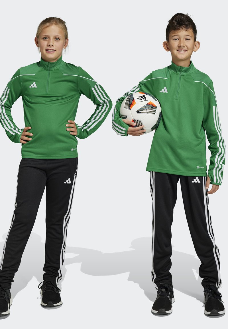 Grüne sportliche Zip-Up-Tops mit weißen Streifen und schwarze Hosen mit weißen Seitenstreifen, getragen von zwei Kindern, von denen eines einen Fußball hält.
