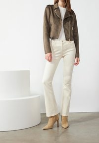 Donna che indossa una giacca corta in suede marrone, una maglietta bianca, jeans beige a vita alta e stivaletti con tacco in suede beige, in piedi all'interno.