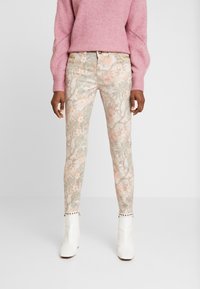 Pantalones ajustados con estampado floral en colores pasteles sobre una base de color beige claro. Presentan accesorios decorativos en la cintura y un detalle de tachuelas en los tobillos.