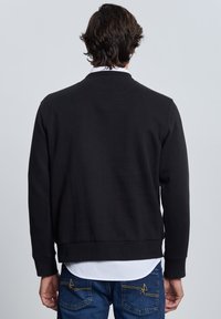 Sudadera negra con cuello redondo y puños acanalados, tela texturizada, usada sobre una camisa blanca con cuello, con un diseño liso en la parte trasera.