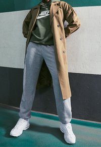 Brun trenchcoat, grön hoodie med vit logotyp, grå sweatpants och vita sneakers. Släta texturer, kontrasterande färger, stilfull lager på lager.