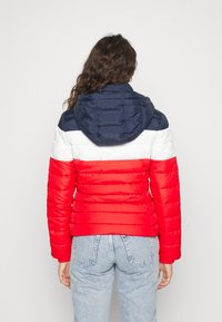 Kvinna med långt, lockigt brunt hår som bär en puffjacka med marinblå, vit och röd horisontell randning och ljusblå jeans, vänd bort.