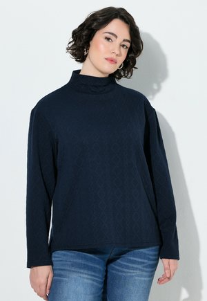 Vrouw met kort krullend donker haar, die een getextureerde donkerblauwe top met lange mouwen en een blauwe spijkerbroek draagt, staand tegen een lichte muur.