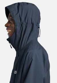 Giacca impermeabile blu marina con cappuccio, dotata di cerniera, coulisse regolabile e una texture leggera e liscia.