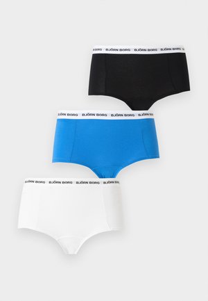 Žena stojaca na boso, oblečená v čiernej podprsenke a čiernych krátkych boxerkách s bielym elastickým pásom na jednoduchom pozadí.
