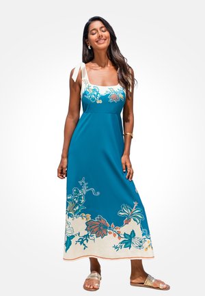 Frau mit langen dunklen Haaren, die ein blau-weißes, geblümtes Maxikleid mit Schulterbändern und goldenen Sandalen trägt, steht mit geschlossenen Augen und lächelt.